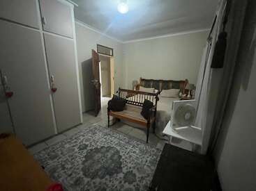 A cozy, dimly-lit bedroom with a large bed, wooden bench, patterned rug, wardrobe, side table, fan, open door, and simple decor. Inviting atmosphere.