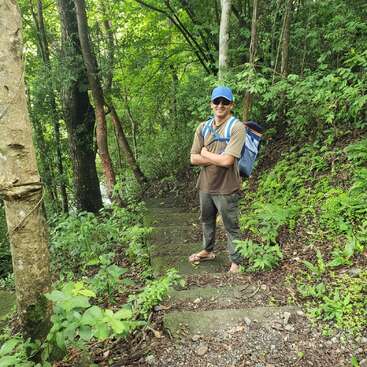 Un hombre con gafas de sol, gorra azul, mochila y chanclas está de pie sobre unos escalones de piedra en un frondoso bosque verde, sonriendo con los brazos cruzados.