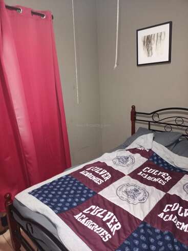 Um pequeno quarto com paredes cinza, uma estrutura de cama de metal preta, cobertor da Culver Academies, cortinas vermelhas, arte na parede emoldurada e cordas penduradas no teto.