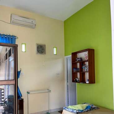Una habitación pequeña y ordenada con una cama individual, una pared decorativa en verde, una estantería empotrada, aire acondicionado, cortinas azules, suelo de baldosas y un mobiliario minimalista que crea un ambiente acogedor.