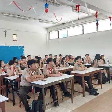 A classroom filled with students in brown uniforms attentively listens to someone at the front. The room is decorated with red and white streamers.