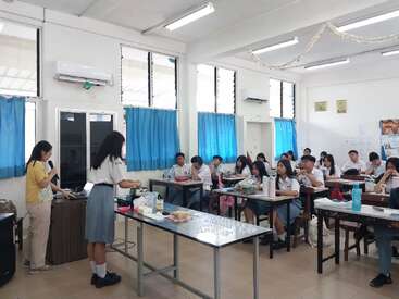 In einem Klassenzimmer mit blauen Vorhängen hält ein Schüler vorne einen Vortrag, während die Mitschüler sitzen und aufmerksam zuhören. Lehrmaterialien und Lebensmittel sind ausgestellt.