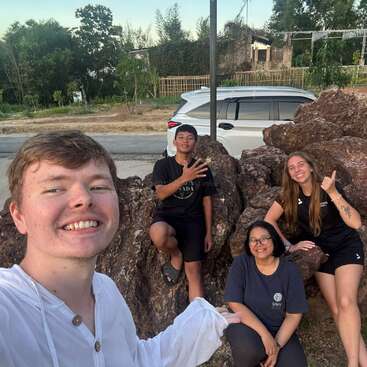 Quatre amis posent joyeusement pour un selfie de groupe en plein air, assis sur de gros rochers. Derrière eux, on aperçoit des arbres, une voiture blanche et un bâtiment partiellement visible. Tout le monde sourit.