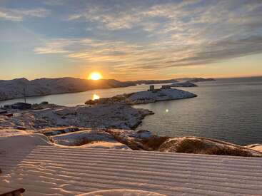 Das Bild zeigt eine ruhige Winterlandschaft mit schneebedeckten Bergen und Inseln vor einem leuchtenden Sonnenuntergangshimmel, mit einem Gewässer und einem kleinen Dorf in der Ferne.