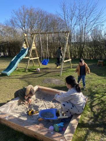 Vier Kinder spielen an einem sonnigen Tag im Freien. Zwei sind in einem Sandkasten mit Spielzeug, während zwei andere in der Nähe einer Schaukel und einer Rutsche spielen. Sie sind von Bäumen umgeben.