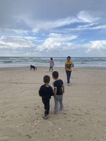 Vier Kinder und ein Hund genießen einen Sandstrand unter einem teilweise bewölkten Himmel. Zwei Jungen gehen zusammen, ein Mädchen steht mit dem Hund, ein anderer Junge lächelt.