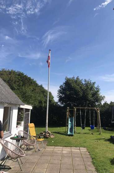 Ein sonniger Hinterhof mit einer kleinen Terrasse, Stühlen, einer dänischen Flagge, üppigem Grün, einer Kinderschaukel mit Rutsche und einem strahlend blauen Himmel.