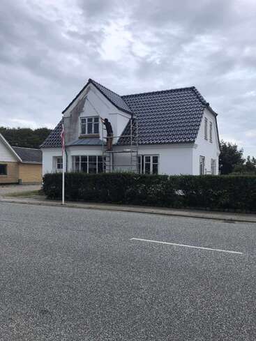 Eine Person steht auf einem Gerüst und streicht die obere Wand eines weißen Hauses mit Ziegeldach, unter einem bewölkten Himmel, mit Blick auf eine ruhige Straße.