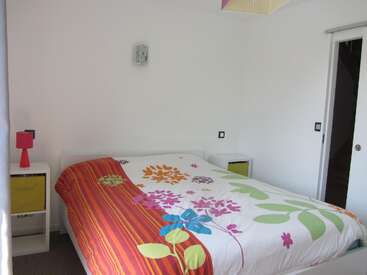 Un dormitorio luminoso y minimalista cuenta con una cama de matrimonio con un colorido edredón floral, dos mesillas de noche blancas, una lámpara roja, paredes blancas y una puerta corredera.