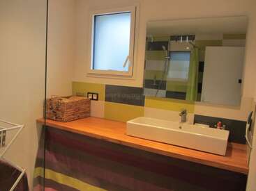 Este cuarto de baño presenta un moderno lavabo rectangular, una encimera de madera, un gran espejo, azulejos a rayas, una pequeña ventana, una cesta de mimbre y una ducha que se refleja en el espejo.