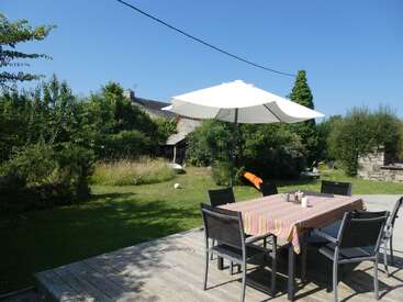 Un soleado patio ajardinado cuenta con una mesa con seis sillas y una sombrilla, rodeado de exuberante vegetación y césped bajo un cielo azul despejado.