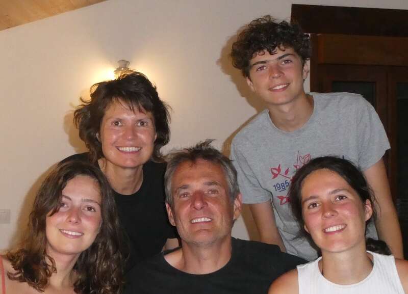 Esta imagen muestra a una sonriente familia de cinco miembros en el interior. Tres adultos jóvenes y dos padres, todos muy juntos. La iluminación cálida, el ambiente relajado y la ropa informal sugieren felicidad.