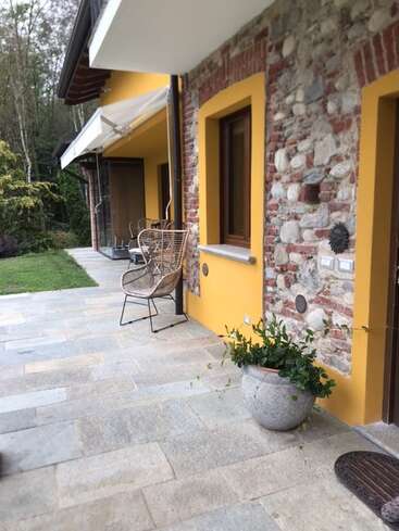 Un charmant patio avec des murs en pierre et jaunes, une chaise en osier, une plante en pot, un sol carrelé et une pelouse verte en bordure d'une zone boisée, dégageant une atmosphère chaleureuse.