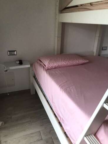 Cette image montre un lit superposé inférieur soigneusement fait avec des draps de lit roses et un oreiller. Une petite étagère blanche contient une montre et un chargeur.