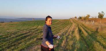 A woman stands in a grassy field at sunset, holding a golf club, looking back and smiling. The landscape is open, peaceful, and surrounded by trees.