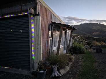 A rustic shed with corrugated iron walls and windows stands at dusk, surrounded by plants, gravel path, and distant hills under a cloudy sky.