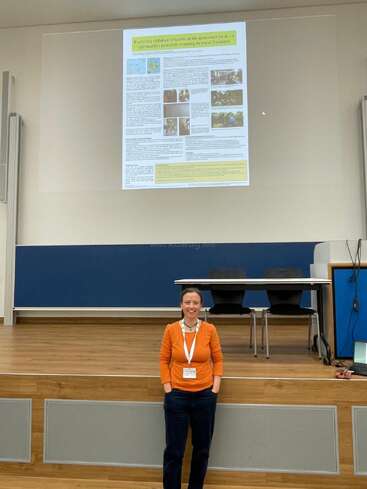 A woman stands smiling in front of a stage, wearing an orange top and name badge. Behind her, a presentation about pediatric training in Vanuatu is projected.