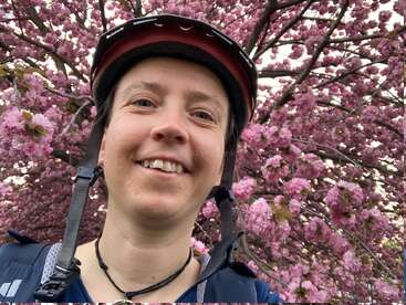 A smiling person wearing a bicycle helmet takes a selfie in front of a beautiful cherry blossom tree filled with vibrant pink flowers in full bloom.