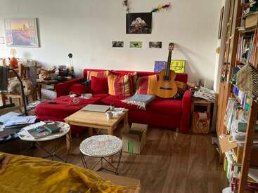 A cozy, cluttered living room with a red couch, colorful pillows, guitar, coffee tables, laptop, books, boxes, and wall art, exuding a creative atmosphere.