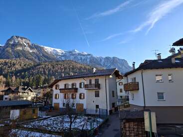 Encantador pueblo alpino con casas tradicionales, enclavado a los pies de majestuosas montañas nevadas. Predominan el cielo azul y despejado, las colinas boscosas y un ambiente tranquilo.