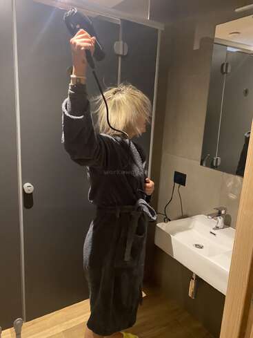 Una persona vestida con un albornoz oscuro se está secando el pelo con un secador en un cuarto de baño moderno, de pie junto a un lavabo y un espejo blancos.