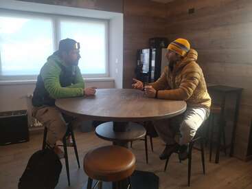 Dos hombres vestidos con ropa de invierno están sentados alrededor de una mesa redonda en una acogedora sala de madera, manteniendo una animada conversación. Se ven una ventana y una máquina expendedora.