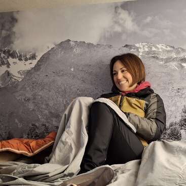 A smiling woman sits comfortably on a bed, wrapped in blankets, wearing a winter jacket. Behind her is a scenic wall mural of snowy mountains and cloudy skies.