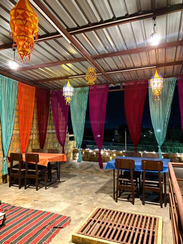 A cozy, colorful terrace setup with draped curtains, vibrant tablecloths, traditional hanging lanterns, wooden chairs, striped rug, and exposed stone walls, glowing under warm lights.