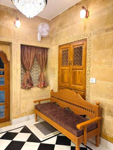 This image shows a cozy room corner with ornate wooden bench, brown cushion, decorative curtains, wall fan, wooden window, chandelier, wall lights, and black-and-white tiled floor.
