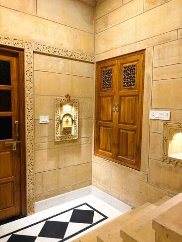 This image shows a beautifully designed interior with carved sandstone walls, wooden doors and window, niche shelves, and intricate traditional Indian architectural details and decor.