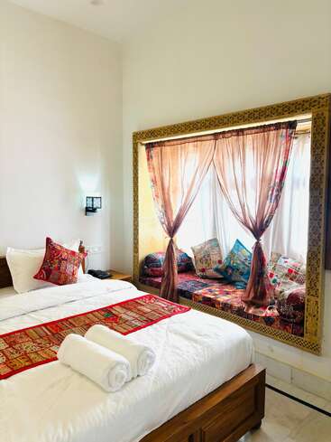 This cozy bedroom features a comfortable bed with white linens, vibrant ethnic-patterned pillows, and a window nook with colorful cushions and sheer curtains. Inviting atmosphere.