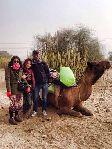 Three people stand together smiling in a sandy, desert-like environment with a seated camel carrying luggage beside them. Tall, green desert plants grow in the background.