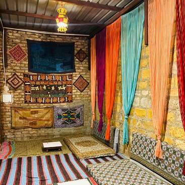 This cozy room features colorful drapes, patterned cushions, and woven rugs. Decorative wall hangings and dim lantern lighting create a warm, traditional Middle Eastern or Indian ambiance.