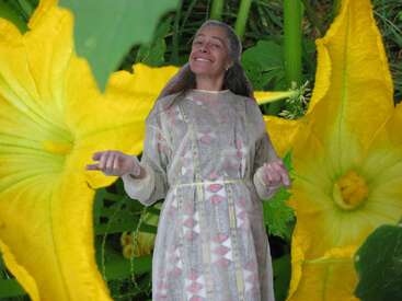 L'image représente une femme vêtue d'une robe blanche, debout au milieu de grandes fleurs jaunes et de feuilles vertes, les mains tendues devant elle.