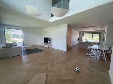 This image shows a spacious, modern living room with wooden herringbone flooring, minimalist furniture, a TV, dining area, large windows, and a soccer ball on the floor.
