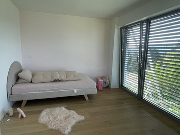 A minimalist bedroom with a single bed, light bedding, wooden floor, fluffy pink rug, large windows with blinds, and a few personal items in the corner.
