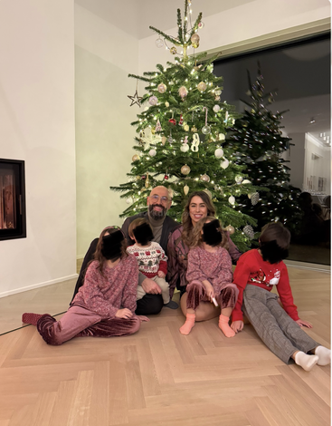 A smiling family of six sits together in front of a beautifully decorated Christmas tree, celebrating the festive season in a cozy, warmly lit living room.