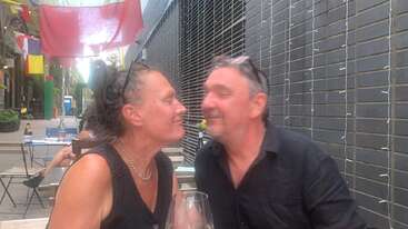 A smiling couple sits closely at an outdoor café, sharing a joyful, intimate moment. Colorful flags hang above, and wine glasses are on the table.