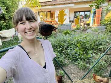 Una mujer sonriente se hace un selfi en su jardín. Una gallina negra se posa en su hombro. Detrás de ella, una acogedora casa y la vegetación crean una escena alegre.