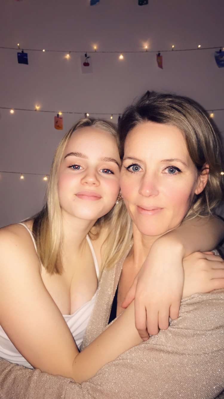 The image shows two women posing for a photo, with one woman\'s arms wrapped around the other\'s shoulders, in front of a wall adorned with string lights.