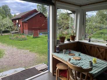 Dieser gemütliche Raum verfügt über einen karierten Tisch, Topfpflanzen und Holzstühle an großen Fenstern, von denen aus man auf einen üppig grünen Garten und eine klassische rote Scheune blickt.