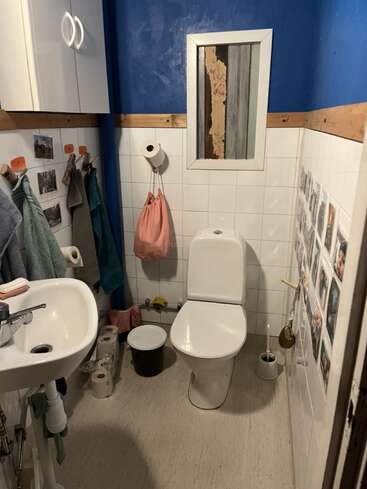Das ist ein kleines Badezimmer mit blauen Wänden, weißen Fliesen, einer Toilette, einem Waschbecken, Handtüchern, Fotos an den Wänden, einem Schrank und mehreren Rollen Toilettenpapier.