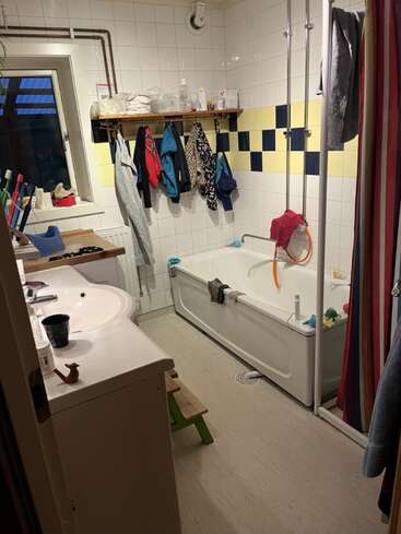 Ein gemütliches Badezimmer mit Badewanne, bunten Handtüchern, einem gestreiften Duschvorhang, aufgehängter Kinderkleidung, Pflegeprodukten, einem Fenster, Ablageflächen und verstreutem Badespielzeug. Eine wohnliche Atmosphäre.