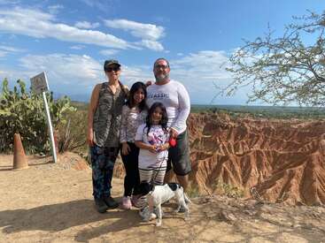 Une famille de quatre personnes souriantes pose avec leur chien devant un panorama spectaculaire composé de canyons désertiques, d'un ciel lumineux, de cactus et d'une végétation clairsemée.