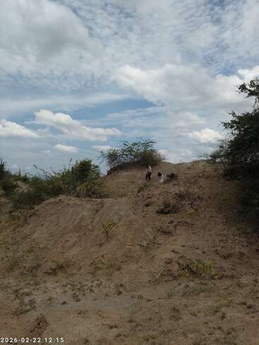 Une colline sablonneuse parsemée d'arbustes sous un ciel nuageux. Deux personnes escaladent la colline. Le paysage semble aride, avec une végétation clairsemée et un temps partiellement ensoleillé.