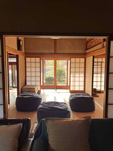 Una serena habitación de estilo japonés con puertas shoji, suelo de tatami, grandes pufs, suave luz solar y vistas al jardín a través de puertas de cristal con marco de madera al fondo.