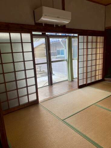 Esta imagen muestra una habitación tradicional japonesa con suelo de tatami, puertas correderas shoji, un aparato de aire acondicionado encima y vistas al exterior a través de puertas de cristal.