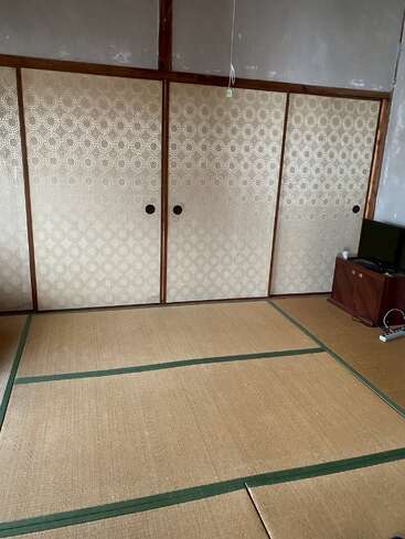 Esta imagen muestra una habitación japonesa tradicional con suelo de tatami, puertas correderas shoji, paredes lisas y un pequeño televisor en un rincón. Decoración minimalista.