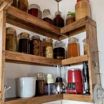 Los estantes de madera de un rincón de la cocina albergan tarros de cristal de conservas y líquidos, junto con electrodomésticos de cocina como una batidora, un robot de cocina y una batidora roja.