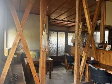 El interior de esta acogedora cafetería presenta vigas de madera, muebles sencillos, plantas colgantes y una luz tenue. Cojines, libros y decoración crean un ambiente cálido, acogedor y relajado.